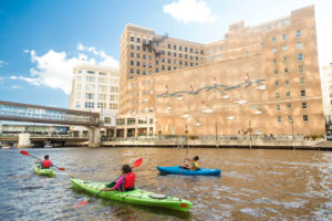 Boaters kayak down the Milwaukee River in green and blue kayaks next to a brown building with a sculpture of waves and fishes along its side.