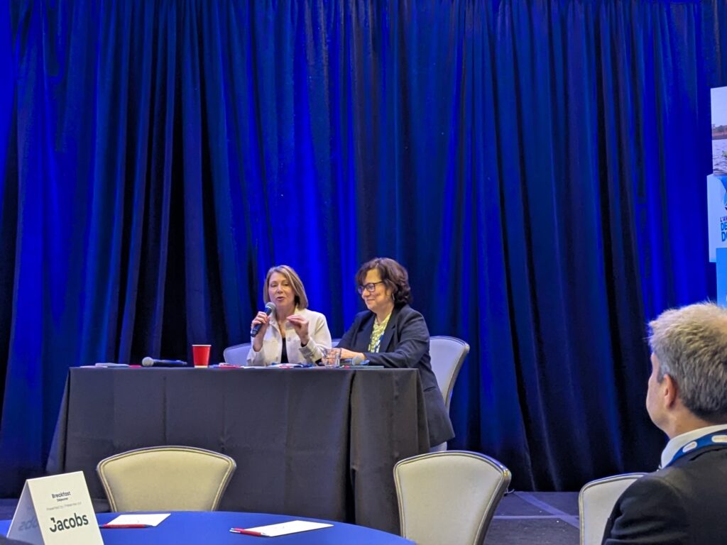 Two women sit at a table on a stage with a blue curtain behind them. One speaks into a microphone.