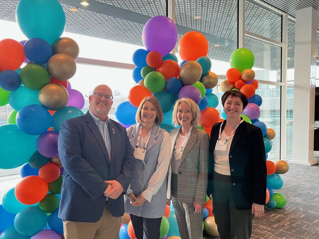 Three woman and one man pose in front of colorful balloons.
