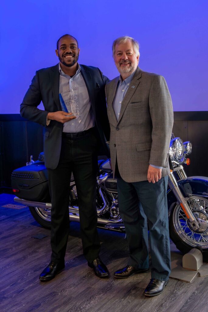 Two men stand in front of a motorcycle on display. One holds a glass award.
