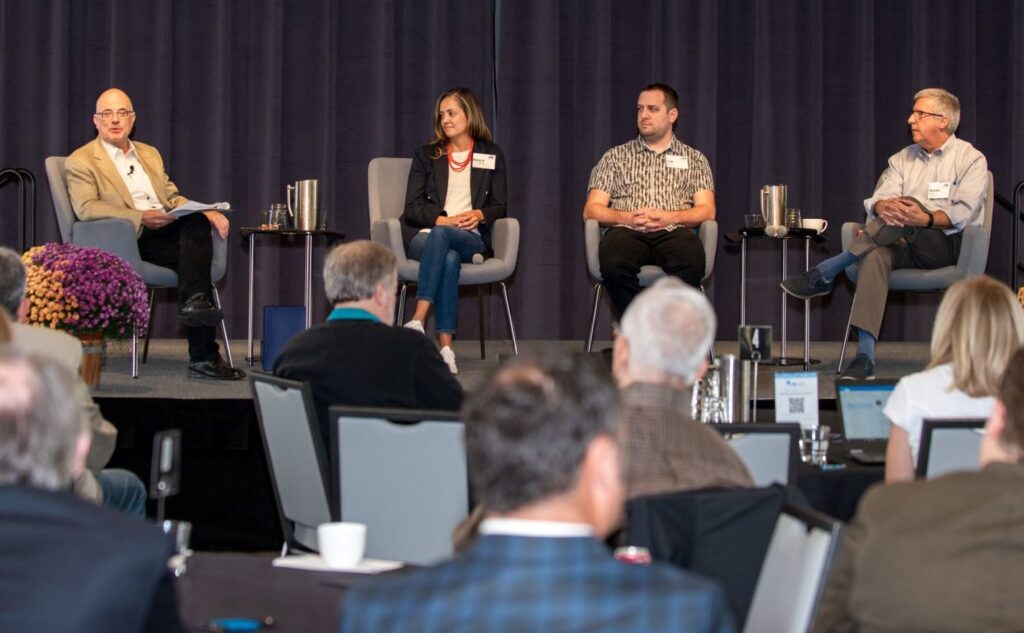Three men and one woman sit on chairs on a stage with flowers in front of them.