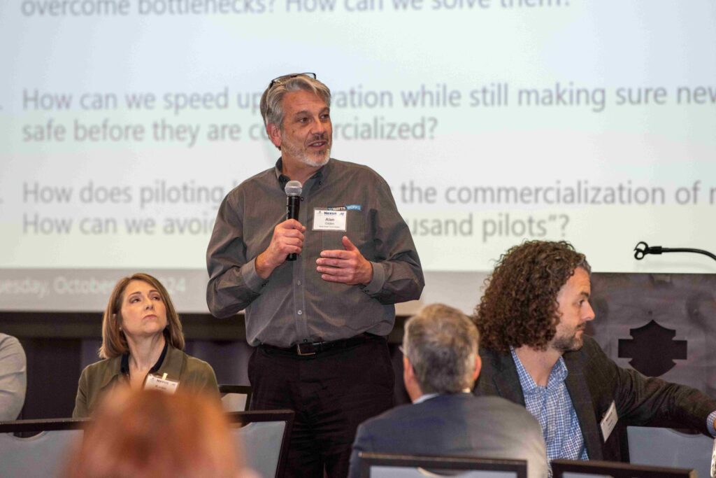Man in gray business shirt stands in front of a large projector screen holding a microphone.