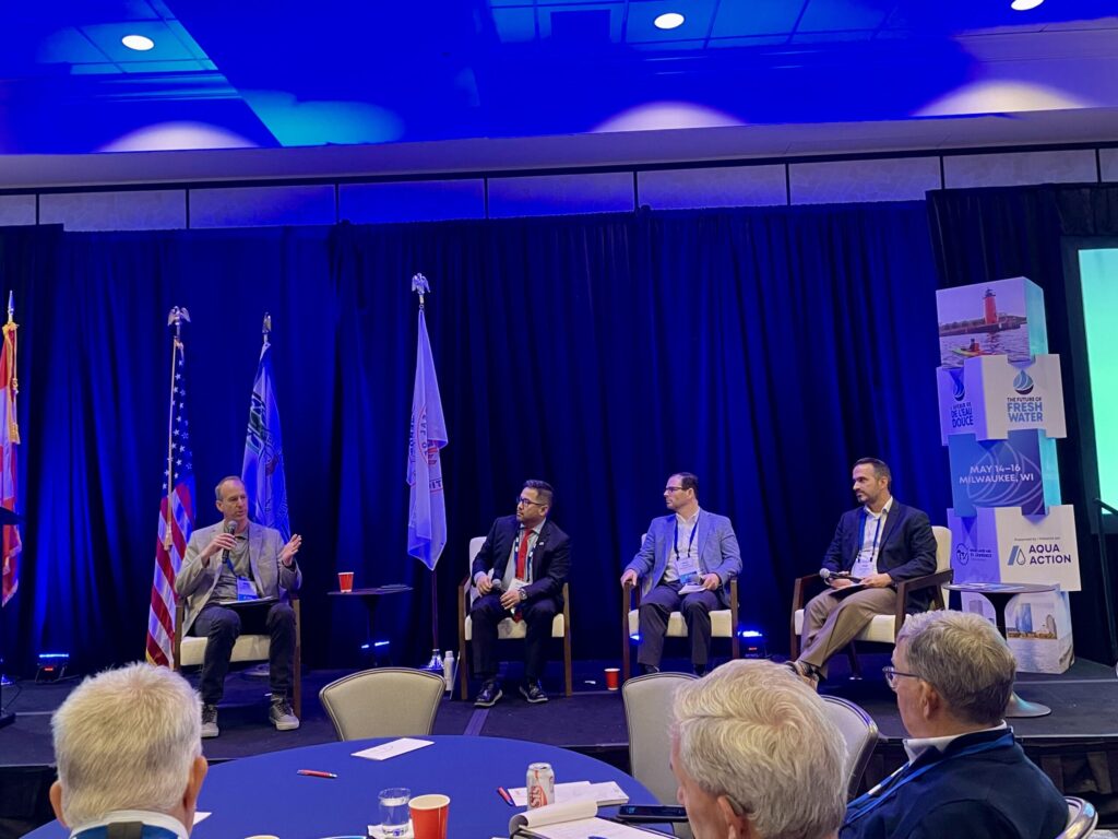 Four men sit on a stage. The one on the left is speaking into a microphone.