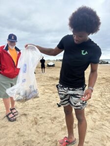 Teen on the beach holds up garbage bag half full of rash while adult looks on behind him.