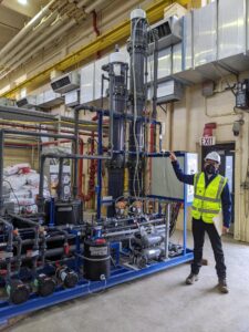 Man in hard hat and yellow vest points to series of pipes with two tall tubes