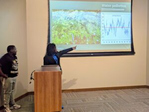 Teen boy in suit jacket stands at a podium and points to a slide with a chart on the screen behind him.