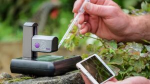 A hand places a drop of water on a small electronic device using a plastic pipette. A second hand is holding a smartphone in front of the device.