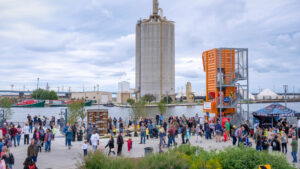 A large group of people gather along the lakefront in front of an orange play structure.