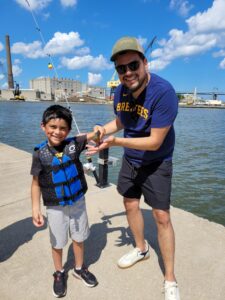 An adult man and a young boy proudly show a fish they have caught standing on a boardwalk next to the water.