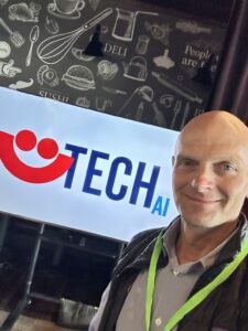 Man smiles in front of banner with red smiley face (Summerfest logo) that says TECH AI.