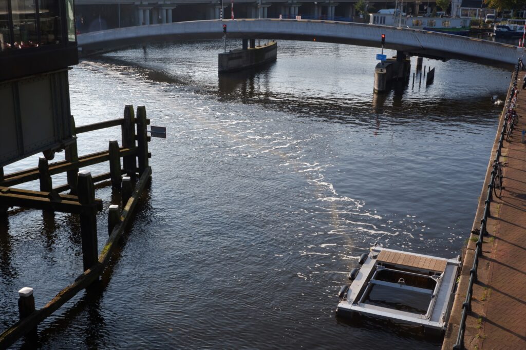 Photo of a river with a bridge across it. A square device on the bank of the river releases bubbles across the river.