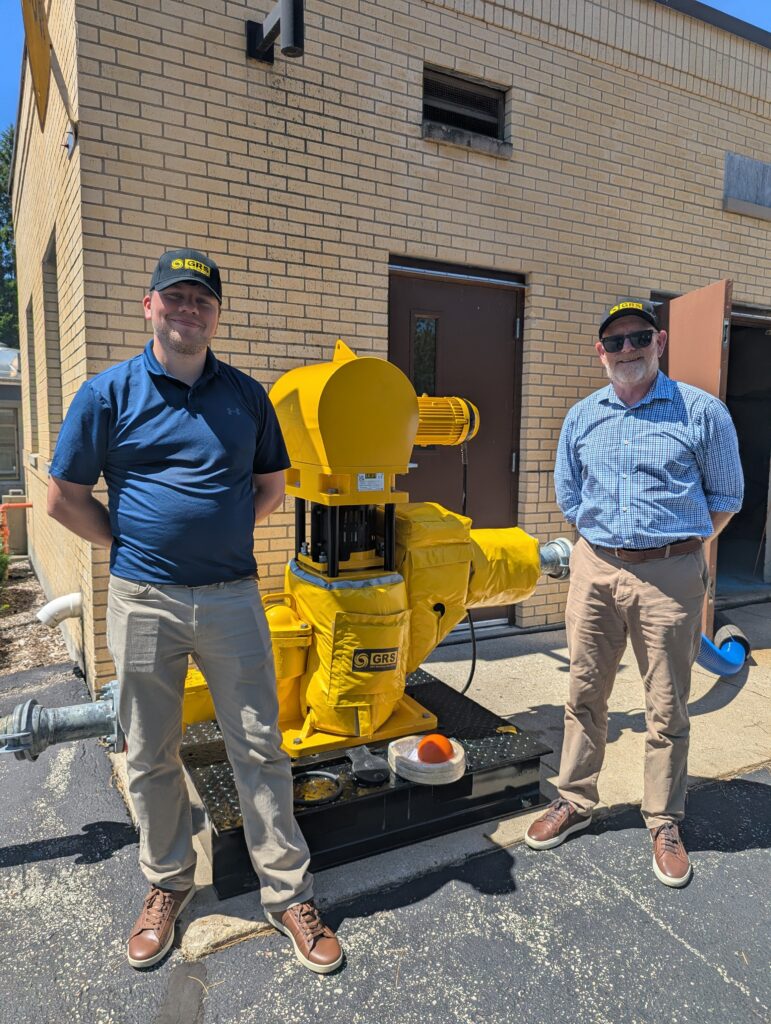 Two men wearing khakis and blue shirts stand next to a 5-foot yellow pump in front of a brick building.