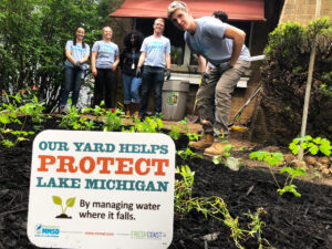 Young adults in matching white T0shirts surround a garden with small green plants and the sign "This yard helps protect Lake Michigan."