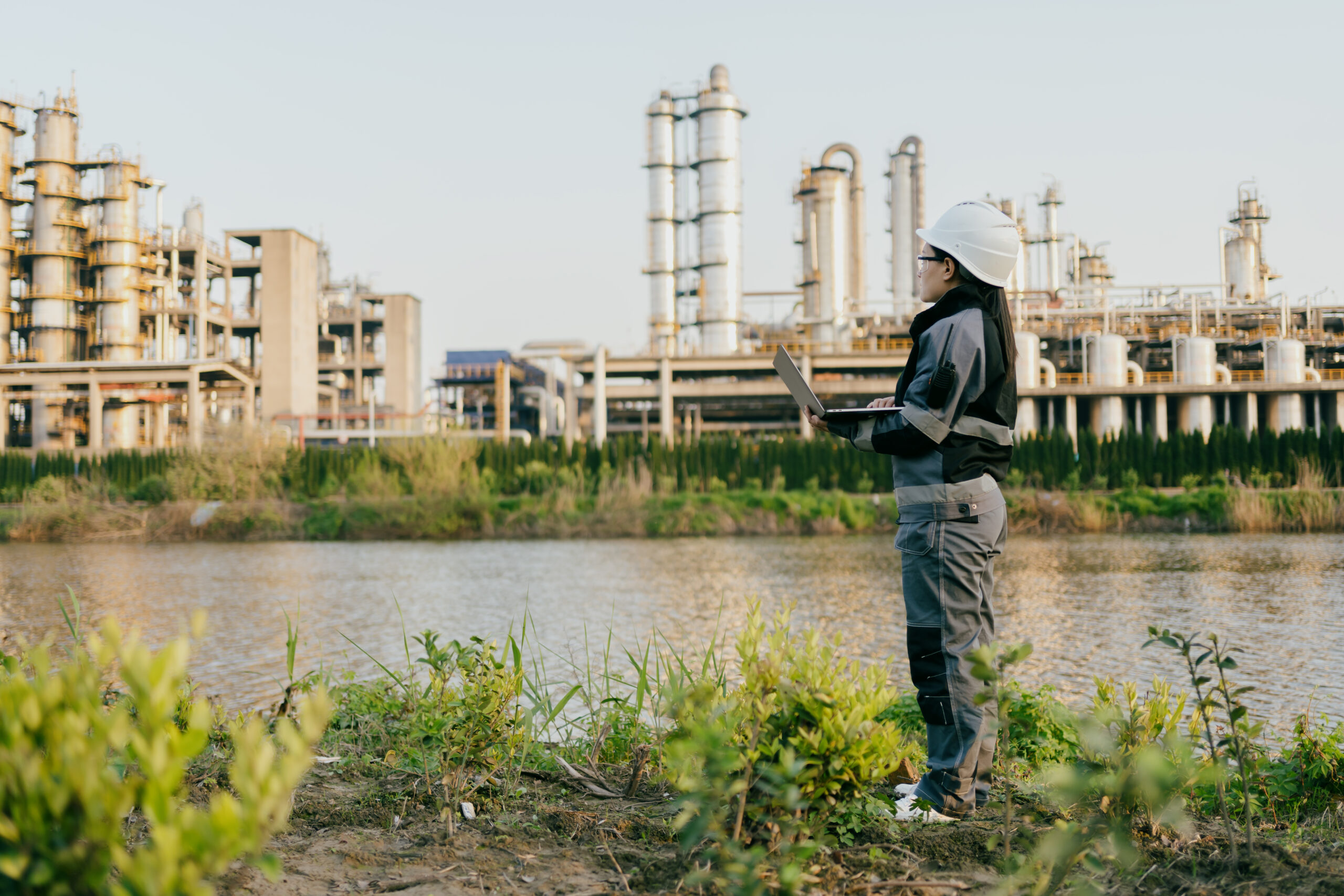 Woman in gray jumpsuit with hard hat and safety glasses holds laptop, looking across a river to an industrial plant.