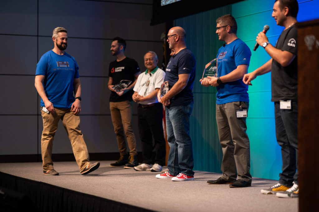 A group of men stand in a line on a stage with a blue background as one man walks in front of them to accept an award.