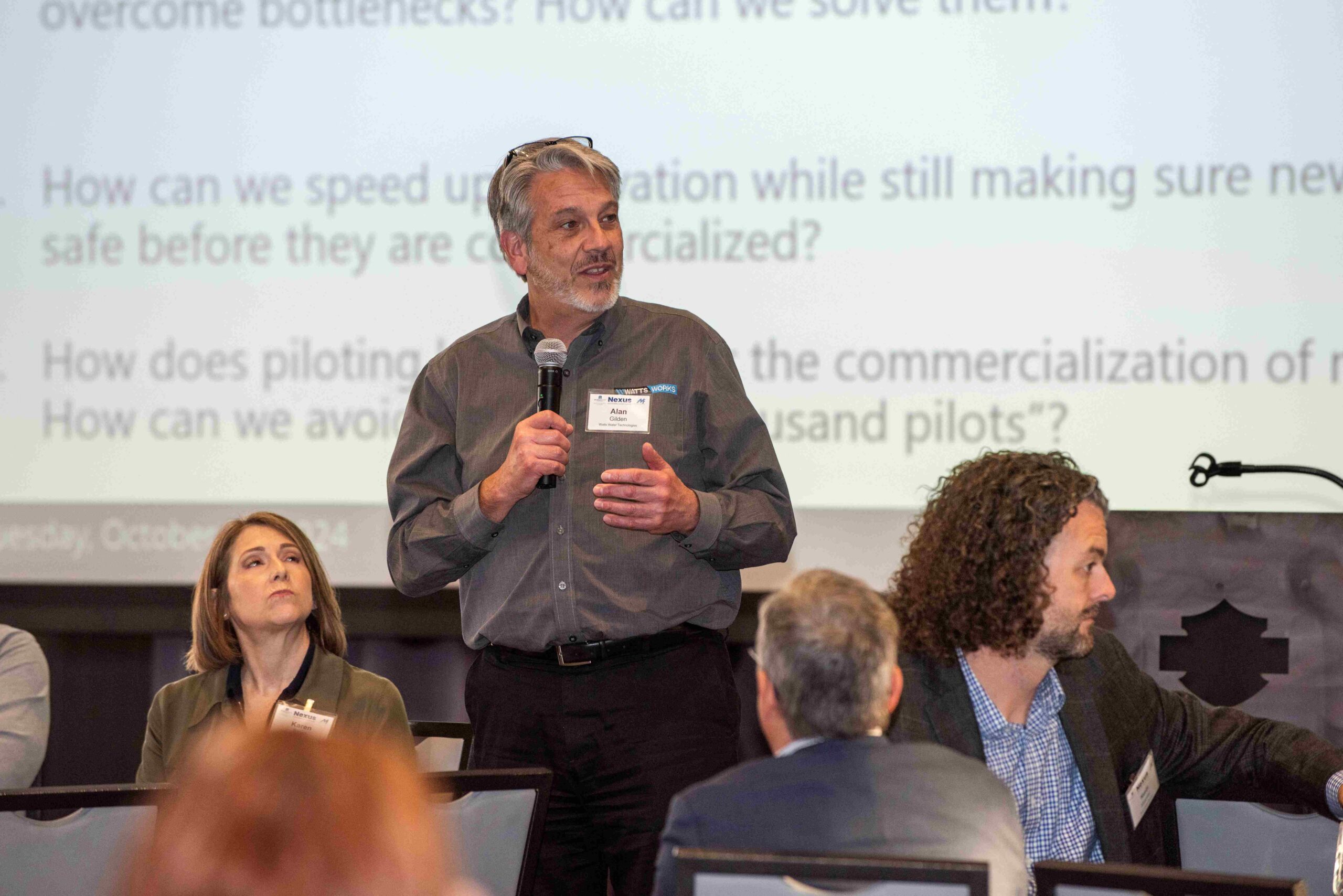 Man in gray business shirt stands in front of a large projector screen holding a microphone.