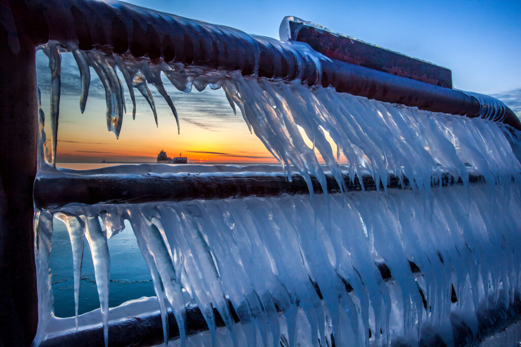 Large icicles hang from a railing, with a view of Lake Michigan  and a lighthouse at sunrise just beyond.