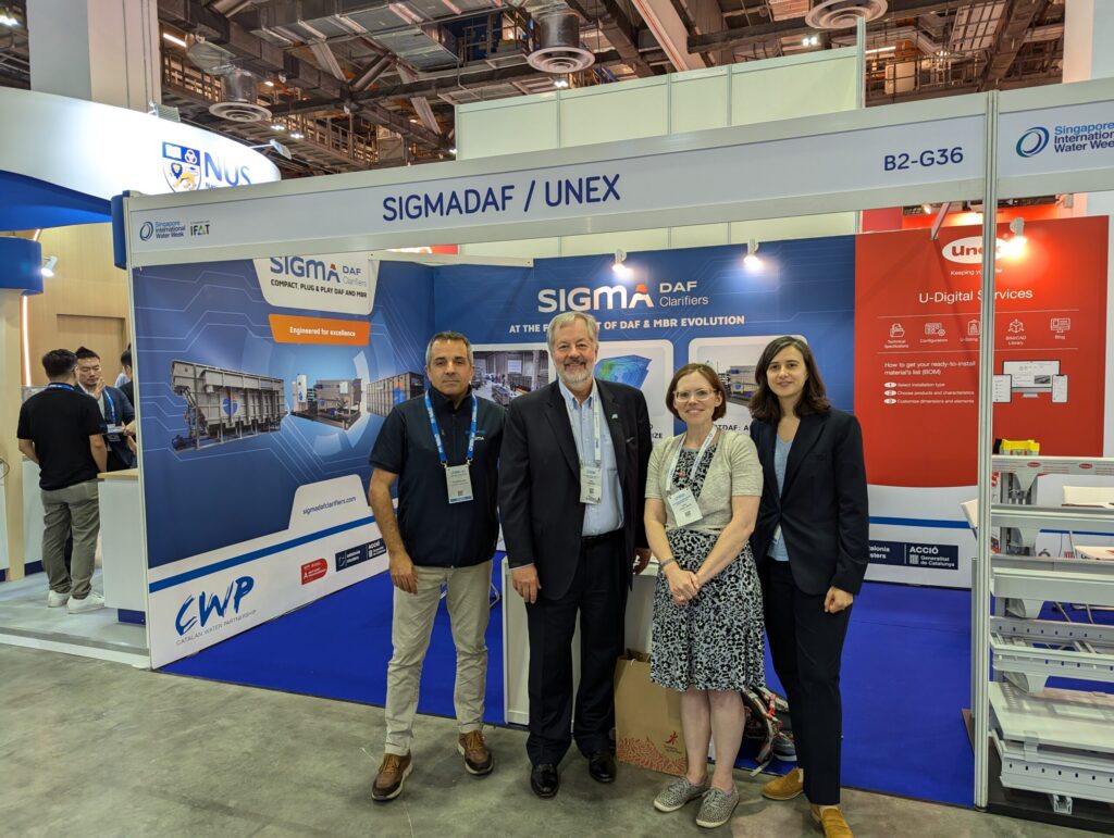 Four people pose for a photo at a booth for the company Sigma DAF at the SIWW exhibit hall.