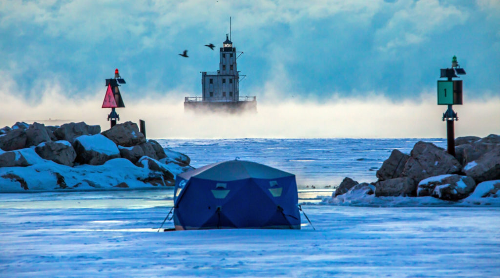ice fishing tent/shanty on the ice with breakers and a lighthouse behind