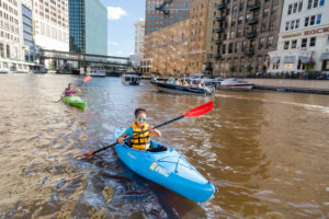 Woman in sunglasses and yellow lifevest paddles a blue kayak down the Milwaukee River with downtown buildings in the background