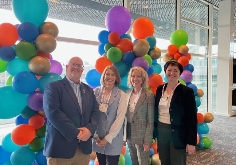 Three woman and one man pose in front of colorful balloons.