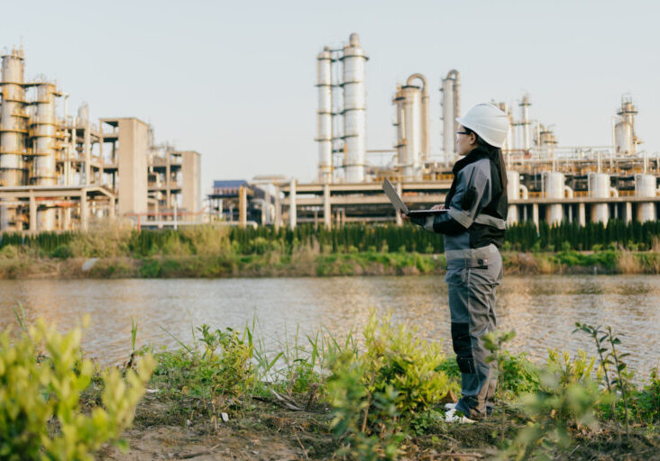 Woman in gray jumpsuit with hard hat and safety glasses holds laptop, looking across a river to an industrial plant.