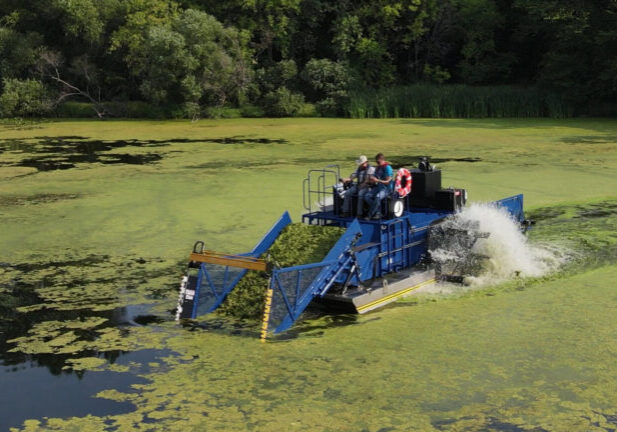 A stationary float with two people seated on top sits on a body of water covered in green algae