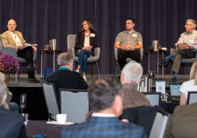 Three men and one woman sit on chairs on a stage with flowers in front of them.
