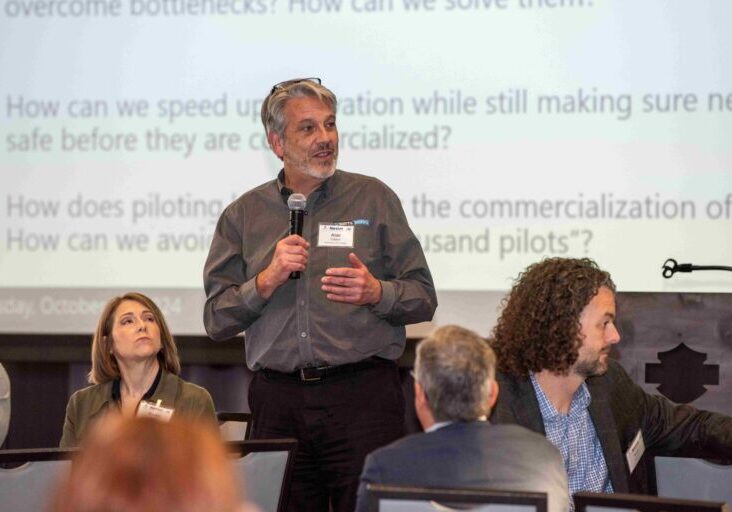 Man in gray business shirt stands in front of a large projector screen holding a microphone.