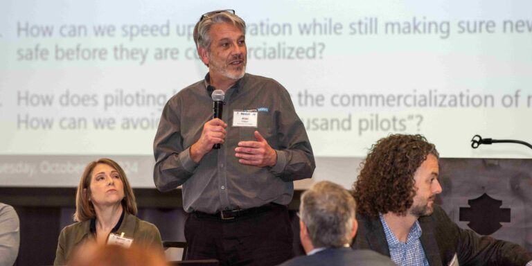 Man in gray business shirt stands in front of a large projector screen holding a microphone.