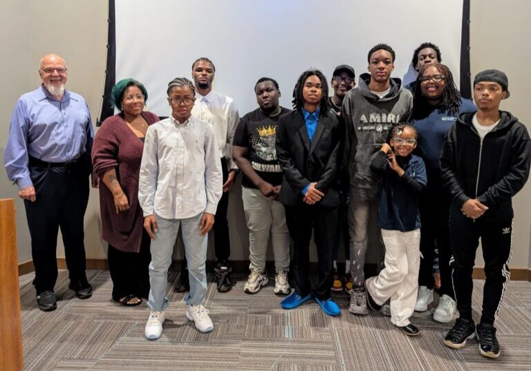 Group of teen boys and several adults spose in front of a projector screen.