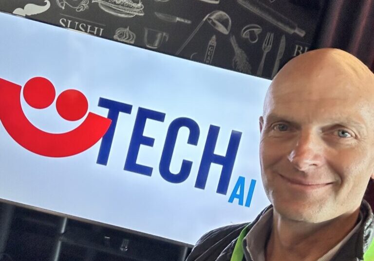 Man smiles in front of banner with red smiley face (Summerfest logo) that says TECH AI.