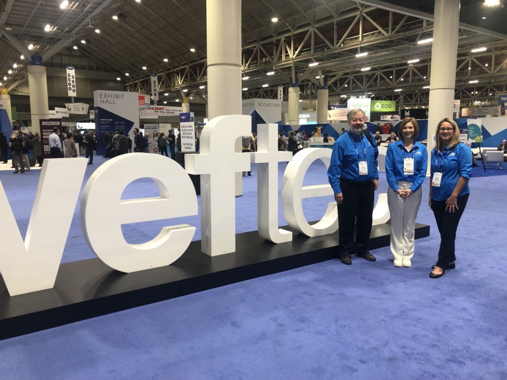 A man and two women in blue shirts pose in front of large letters that spell "WEFTEC"