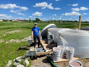 Young woman in blue shirt and baseball cap stands next to large water cylinders in a field.
